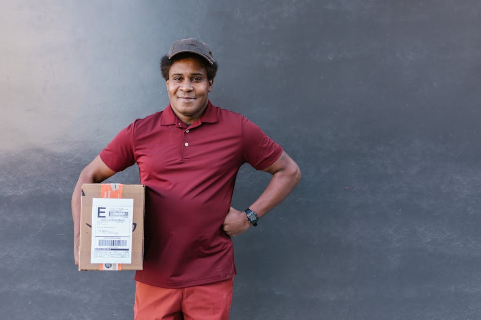 A cheerful male delivery person dressed in a maroon polo shirt, holding a small parcel with an airmail label in his left hand. He stands against a plain, dark grey wall, smiling confidently at the cam