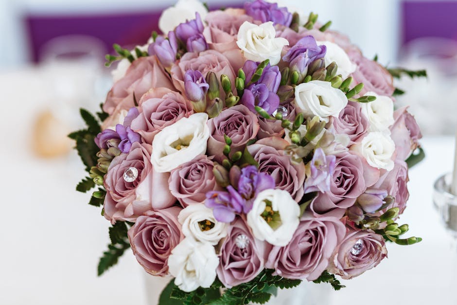 A close-up of a lush, round floral bouquet featuring soft pink roses, white lisianthus, and light purple freesia, accented with small green leaves and clear decorative gems. The flowers are arranged t